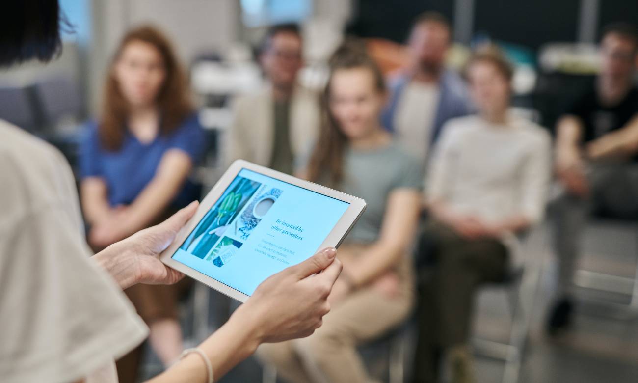 Picture of a group of people listening to a speaker holding a tablet. Photo by fauxels from Pexels.